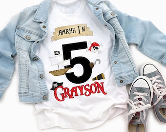 May include: A white t-shirt featuring a large black number 5, the name Grayson in red, and pirate-themed graphics including a ship, a skull with an eyepatch, a hook, a telescope, and a pirate hat, with the text AARHH I'M above the number 5.