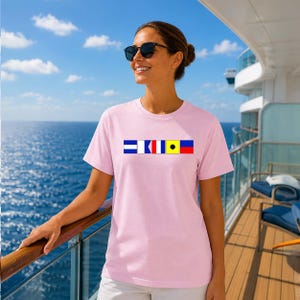 May include: A pale pink t-shirt displaying a line of nautical signal flags in blue, white, red, and yellow. The shirt is worn on a cruise ship deck, with the sea and sky visible in the background.