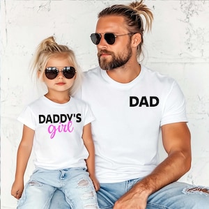 May include: Two white t-shirts with black text. One says "DAD" and the other says "DADDY'S girl" in black and pink. The shirts are worn by a man and a young girl, both wearing sunglasses.
