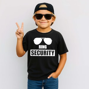 May include: A black short-sleeved t-shirt featuring a white graphic of aviator sunglasses above the text RING SECURITY in a bold, sans-serif font. The design is centred on the chest of the shirt, which is worn with blue denim jeans.