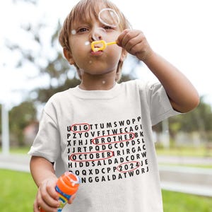 May include: A white short-sleeved t-shirt featuring a black word search puzzle graphic. The words BIG, BROTHER, OCTOBER, and 2024 are circled in red ink. The shirt has a crew neckline and a standard fit, displaying the text clearly on the front.