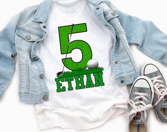 May include: A white t-shirt featuring a large green number five with a distressed texture, a golf club, a golf ball, and a patch of green grass. The name ETHAN is printed in green capital letters below the grass graphic, accompanied by a light blue denim jacket and a pair of gray canvas sneakers.