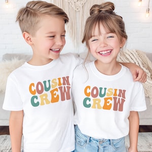 May include: Two white short-sleeve t-shirts featuring the text COUSIN CREW printed in a wavy, retro-style font. The letters are colored in shades of orange, yellow, teal, and muted red. The shirts have a classic crew neckline and straight hem.