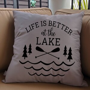 May include: A gray throw pillow with black text that reads "Life is better at the lake". The text is surrounded by a graphic of two crossed paddles, waves, and pine trees.