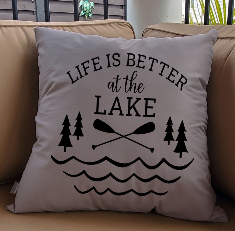 May include: A gray throw pillow with black text that reads "Life is better at the lake". The text is surrounded by a graphic of two crossed paddles, waves, and pine trees.