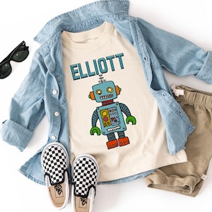 May include: A beige t-shirt with a blue robot graphic and the name "ELLIOTT" printed in black. The shirt is paired with a light blue denim jacket and khaki shorts.