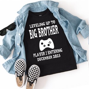 May include: Black t-shirt with white text that reads "Leveling Up To Big Brother" and "Player 2 Entering December 2025" with a white video game controller graphic. The shirt is paired with a denim shirt, black sunglasses, and black and white checkered slip-on shoes.
