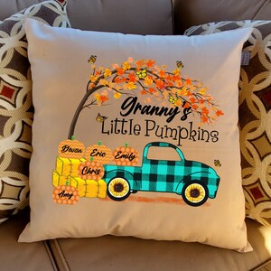 May include: A tan throw pillow with a fall-themed design. The pillow features a turquoise truck with a black and white plaid design, carrying pumpkins with names on them. The truck is driving under a tree with orange and yellow leaves and butterflies. The text "Granny's Little Pumpkins" is printed on the pillow.