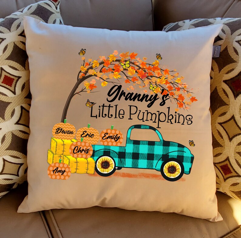 May include: A tan throw pillow with a fall-themed design. The pillow features a turquoise truck with a black and white plaid design, carrying pumpkins with names on them. The truck is driving under a tree with orange and yellow leaves and butterflies. The text "Granny's Little Pumpkins" is printed on the pillow.