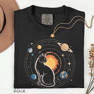 May include: A black t-shirt featuring a graphic print of a cat sitting in front of a large orange sun, surrounded by orbiting planets and stars. The shirt has a Comfort Colors label at the neck and is paired with a gold-toned star pendant necklace.