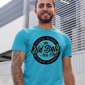 May include: A bright turquoise short-sleeve t-shirt featuring a circular black graphic with yellow accents. The text reads: Official Member The Old Balls Club est. 1976 50 Years of Awesome. The design is centred on the chest of the garment.