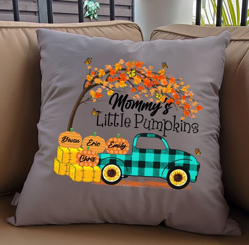 May include: Gray throw pillow with a design of a teal and black plaid pickup truck carrying pumpkins labeled Devon, Eric, Emily, and Chris. The truck is driving under a tree with orange leaves and butterflies. The text "Mommy's Little Pumpkins" is above the truck.