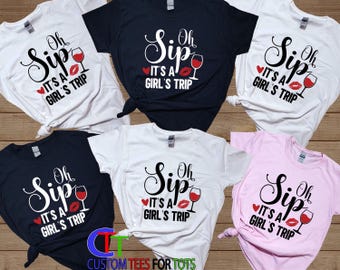 May include: A collection of six short-sleeved t-shirts in white, black, and light pink. Each shirt features the black text 'Oh Sip IT'S A GIRL'S TRIP' with a red heart, a red lip print, and a graphic of a wine glass filled with red wine.
