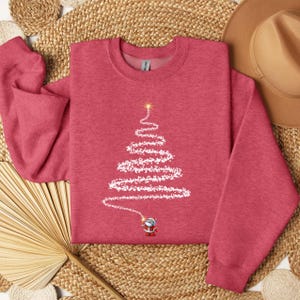 Peut inclure: Un sweat-shirt à manches longues rouge chiné orné d'un motif représentant un petit personnage de Père Noël tenant une baguette lumineuse qui dessine une forme de sapin de Noël blanc et scintillant, surmontée d'une étoile jaune brillante.