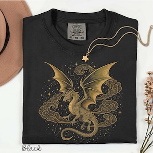 May include: Black t-shirt featuring a gold dragon design with outstretched wings, set against a backdrop of clouds and stars. The shirt has a crew neck and a "COMFORT COLORS" tag. A gold star pendant necklace is on the shirt. The word "Black" is printed at the bottom.