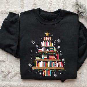 May include: A black long-sleeve sweatshirt featuring a graphic print of a Christmas tree constructed from stacked books on wooden shelves. The design includes a gold star topper, colorful string lights, a black cat, and white snowflakes.