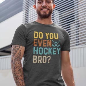 Peut inclure: T-shirt gris foncé avec l'inscription "DO YOU EVEN HOCKEY BRO ?" en jaune, orange, turquoise et beige. Le motif comprend des crosses de hockey. Un vêtement décontracté.
