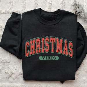 May include: A black crewneck sweatshirt featuring the text CHRISTMAS VIBES in a distressed, collegiate-style font. The word CHRISTMAS is arched in red with a dark green outline, while the word VIBES is in a solid dark green rectangular box.