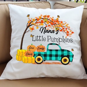 May include: White pillow with a fall-themed design featuring a truck carrying pumpkins labeled "Eric", "Emily", and "Chris". The truck is teal and black plaid with sunflowers on the wheels. The text "Nana's Little Pumpkins" is above the truck.