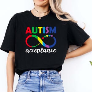 May include: A black short-sleeved t-shirt featuring the text AUTISM in multi-colored block letters above a rainbow-colored infinity symbol. A small row of colorful hearts sits above the symbol, with the word acceptance in white script below.