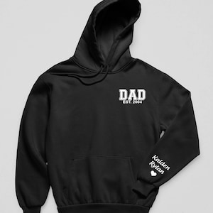 May include: A black hooded sweatshirt featuring white text on the chest that reads DAD EST. 2004 in a collegiate font. The left sleeve displays the names Kaiden and Rylan in a white script font, followed by a small white heart icon below the names.