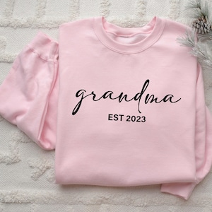 May include: A light pink long-sleeve sweatshirt featuring the word grandma in a black cursive script font, with the text EST 2023 printed in a simple black sans-serif font directly underneath the cursive lettering on the front of the garment.