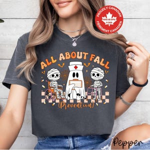 May include: A charcoal grey t-shirt featuring a graphic with the text ALL ABOUT FALL PREVENTION. The design includes two mummies, one in a wheelchair and one with a walker, alongside a ghost nurse holding a clipboard, accented by bats and spiderwebs.