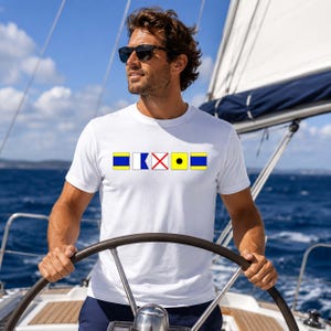 May include: White t-shirt with nautical signal flags in blue, yellow, and red. A person wearing sunglasses is on a sailboat, holding the wheel. The background shows a blue sky and sea. The shirt design is the focal point.