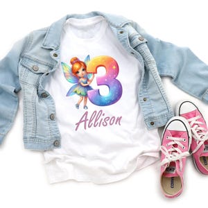 May include: White t-shirt with a fairy graphic holding a number 3. The number 3 is a gradient of colours with a starry design. The name "Allison" is written in a cursive font. A light blue denim jacket and pink trainers are also in the image.
