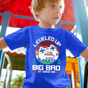 May include: A blue t-shirt with a retro video game controller graphic and the text "LEVELED UP TO BIG BRO EST. FEBRUARY 2026".