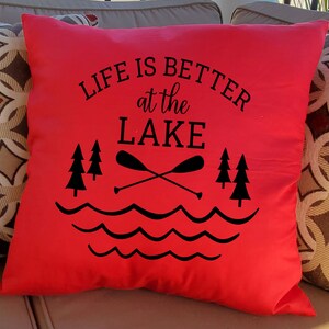 May include: Red throw pillow with black text that reads "Life is better at the lake". The text is surrounded by a graphic of two crossed paddles, three pine trees, and wavy lines representing water.