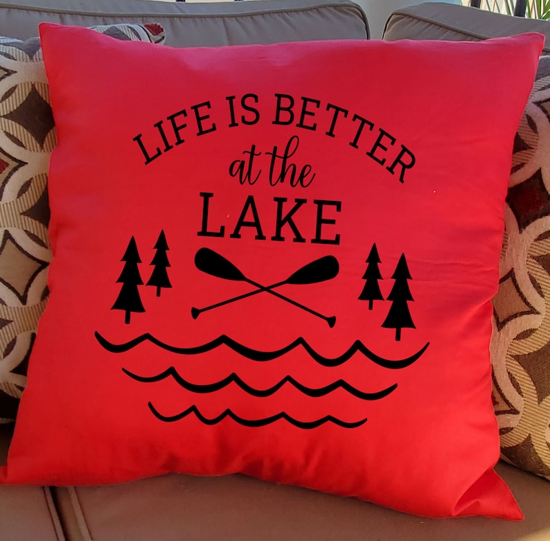 May include: Red throw pillow with black text that reads "Life is better at the lake". The text is surrounded by a graphic of two crossed paddles, three pine trees, and wavy lines representing water.