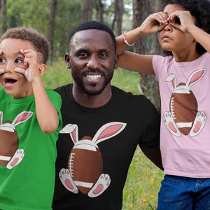 May include: A green t-shirt, a black t-shirt, and a light pink t-shirt, each featuring a graphic of a brown American football with white bunny ears and white bunny feet with pink pads, positioned centrally on the front of the garments.