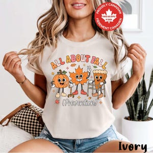 Peut inclure: Un t-shirt ivoire avec un graphique représentant une citrouille, une feuille d'érable et une part de tarte animées. Le texte indique ALL ABOUT FALL (Prevention). La citrouille utilise une canne, la feuille fait un signe de paix et la tarte utilise un déambulateur. Un badge rouge indique PROUDLY CANADIAN.
