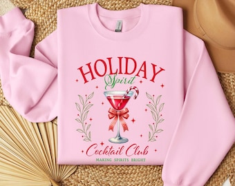 Puede incluir: Una sudadera de cuello redondo rosa claro con un gráfico de un cóctel rojo en una copa con un bastón de caramelo, un lazo rojo y detalles de hojas verdes. El texto dice "HOLIDAY Spirit Cocktail Club" y "MAKING SPIRITS BRIGHT" en rojo y verde.