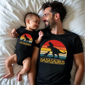 May include: A black baby bodysuit and a black t-shirt featuring a retro sunset graphic with silhouettes of two dinosaurs. The bodysuit has the text JACOBSAURUS and the t-shirt has the text DADASAURUS printed in yellow below the dinosaur graphic.