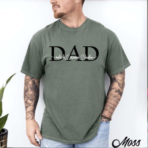 May include: A moss green short-sleeve t-shirt featuring the word DAD in large black capital letters. The names malachi, arianna, and kaiden are written in a white cursive script across the centre of the letters, separated by small heart icons.