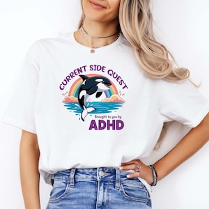 May include: A white t-shirt featuring a graphic of a jumping orca whale, a rainbow, and blue water. The text reads "CURRENT SIDE QUEST" in an arc above the whale and "Brought to you by ADHD" in purple letters below the graphic.