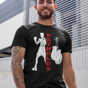 Peut inclure: Un t-shirt noir à manches courtes avec un motif blanc vieilli représentant un boxeur en position de combat, une paire de gants de boxe suspendus et le texte rouge REYNOLDS imprimé verticalement entre le boxeur et les gants.