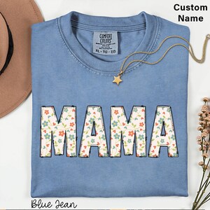 May include: A blue jean coloured t-shirt with a crew neckline and a Comfort Colors label. The front features the word MAMA in large block letters filled with a floral pattern of small blue and orange flowers and green vines. A gold star necklace is included.