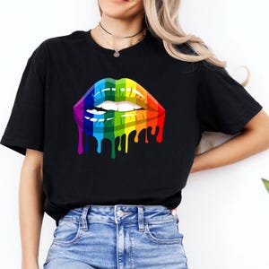 May include: A black t-shirt featuring a graphic print of lips coloured in a rainbow gradient. The lips are depicted with a dripping effect at the bottom, showing white teeth biting the lower lip. The design is centred on the chest of the garment.