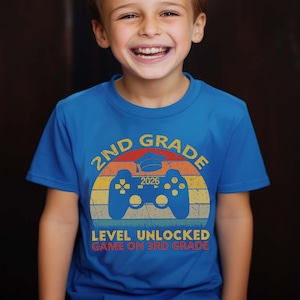 May include: A blue t-shirt featuring a retro-style video game controller graphic. The text on the shirt reads "2nd Grade 2026 Level Unlocked Game On 3rd Grade".