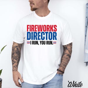 Peut inclure: Un t-shirt blanc à manches courtes avec le texte rouge FIREWORKS et le texte bleu DIRECTOR, suivi du texte noir I RUN, YOU RUN. Le motif comprend de petites rayures horizontales rouges et bleues de chaque côté de la ligne de texte.
