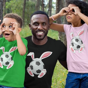 May include: A green t-shirt, a black t-shirt, and a light pink t-shirt, each featuring a graphic of a football with white and pink bunny ears and feet attached to the sides, creating a cohesive set of shirts with a playful animal and sports theme.