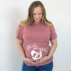 May include: A mauve short-sleeve t-shirt featuring the white text "Turkey Smuggler" above a graphic of a turkey silhouette that incorporates baby footprints into its body design, centered on the front of the garment.