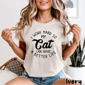 May include: An ivory short-sleeved t-shirt featuring black text that reads I WORK HARD SO MY Cat CAN HAVE A BETTER LIFE. The shirt has a crew neckline and a standard fit, showcasing a clear graphic print centred on the chest area of the garment.