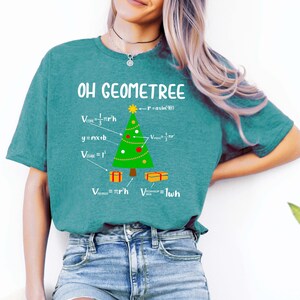 May include: A teal blue t-shirt featuring the text "OH GEOMETREE" and a Christmas tree made of geometric shapes and formulas. The tree is decorated with red and yellow baubles. The text below the tree includes formulas for the volume of a cone, a cube, a cylinder, and a rectangular prism.