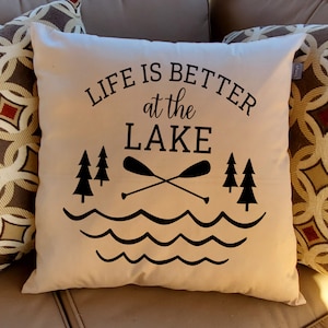 May include: A white throw pillow with black text that reads "Life is better at the lake". The text is surrounded by a graphic of two trees, two crossed paddles, and wavy lines representing water.