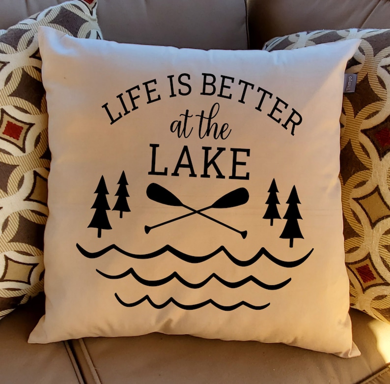 May include: A white throw pillow with black text that reads "Life is better at the lake". The text is surrounded by a graphic of two trees, two crossed paddles, and wavy lines representing water.