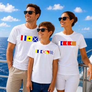 May include: Three individuals wearing white t-shirts adorned with nautical flag designs. The shirts display colourful rectangular patterns. They are wearing sunglasses, standing on a boat with a blue sea and sky in the background.